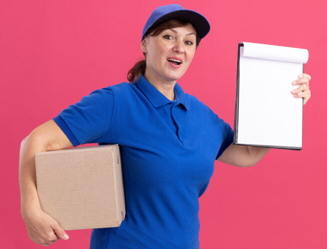 正面中年女送货员穿着蓝色制服 戴着帽子 手里拿着一个纸板箱 上面放着空白页的剪贴板 看着前面 站在粉色的墙上 开心而积极地站着递送中间盒子