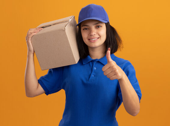 女孩身穿蓝色制服 头戴鸭舌帽 肩上扛着纸板箱 微笑着竖起大拇指 站在橙色的墙上帽子接生拇指
