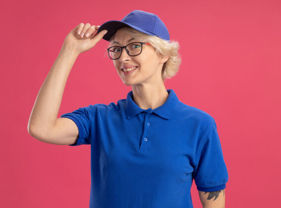 微笑穿着蓝色制服 戴着帽子的快乐的年轻女送货员站在粉红色的墙上 看上去自信满满 戴着帽子微笑着触摸制服女人