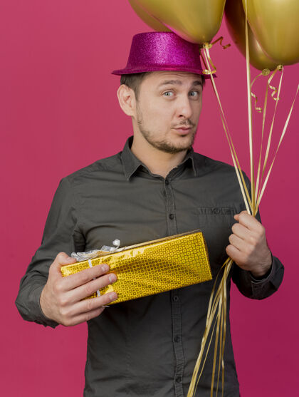 年轻人给人印象深刻的年轻人戴着粉红色的帽子拿着礼品盒气球孤立在粉红色的背景礼物盒子气球