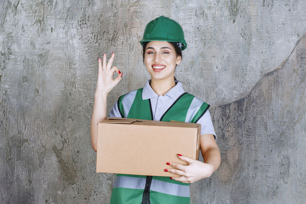积极戴着绿色头盔的女工程师拿着一个纸板箱 展示着满意的标志姿势发货工人