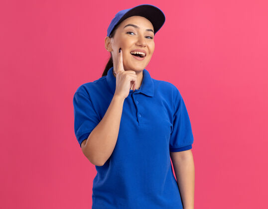 欢呼身穿蓝色制服 头戴帽子的年轻送货员站在粉红色的墙上 面带微笑 开心 积极地看着前面女人帽子交货