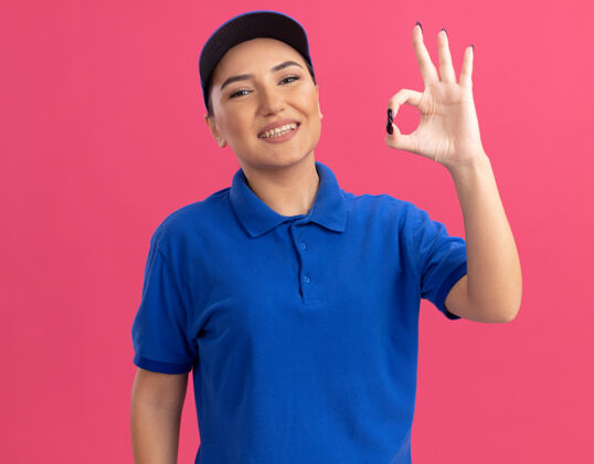 自信身穿蓝色制服 头戴鸭舌帽的年轻女送货员站在粉红色的墙上 自信地微笑着展示着ok标志站立好的制服