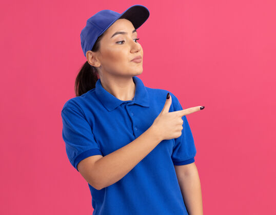 旁边身穿蓝色制服 头戴鸭舌帽的年轻女送货员一边看着 一边自信地用食指指着站在粉色墙上的一边指向立场女人