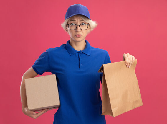 交货身穿蓝色制服 戴着帽子的年轻送货员 手里拿着纸包和纸箱 脸上带着悲伤的表情 站在粉色的墙上悲伤脸包装