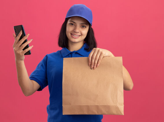 脸身穿蓝色制服 头戴鸭舌帽 手持纸包和智能手机 面带微笑的年轻送货女孩站在粉色的墙上年轻微笑女孩