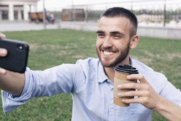 肖像微笑的男人拿着一次性咖啡杯在公园自拍抱着自拍快乐