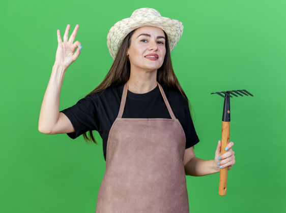 帽子微笑着美丽的园丁女孩穿着制服 戴着园艺帽 拿着耙子 在绿色的背景上表现出很好的姿态显示耙手势