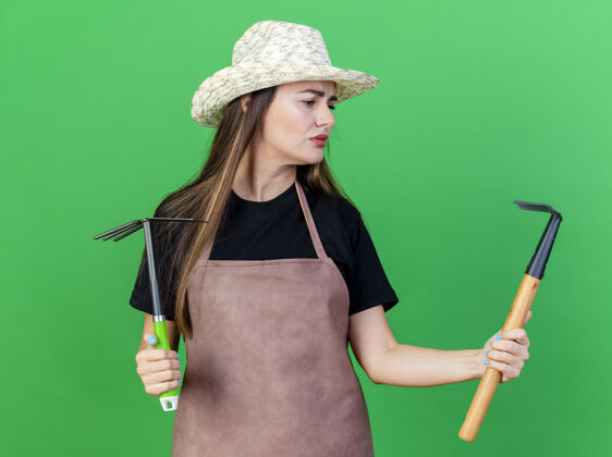 持有迷茫美丽的园丁女孩穿着制服戴着园艺帽拿着锄头耙子看着耙子在她的手上孤立的绿色背景园艺耙子帽子