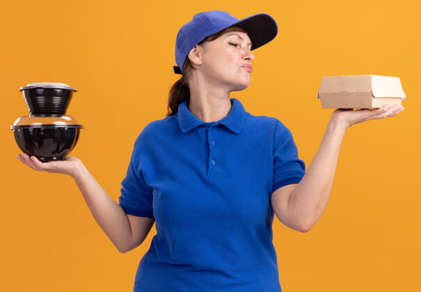 制服身穿蓝色制服 戴着帽子 拿着食品包的中年送货妇女站在橙色的墙上 看上去自信而快乐食物站着自信