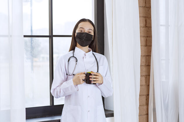 感冒医生带着听诊器和黑色口罩 在休息时端着一杯黑色饮料 暖手治疗医学从业者