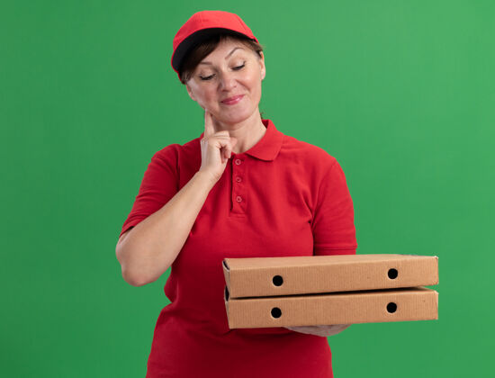 帽子身穿红色制服 头戴鸭舌帽的中年女送货员手持披萨盒 面带微笑地看着披萨盒 站在绿色的墙上 快乐而积极正面快乐站着