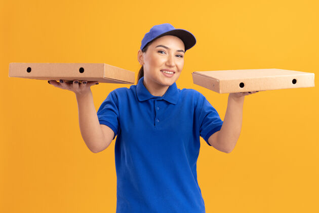 女人身着蓝色制服 头戴鸭舌帽 手持披萨盒 站在橙色墙壁上 面带微笑 快乐地看着前方的年轻送货员年轻微笑快乐