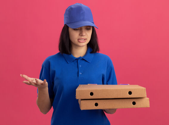 拿着穿着蓝色制服的年轻送货女孩拿着披萨盒站在粉色的墙上困惑地看着它们困惑年轻披萨