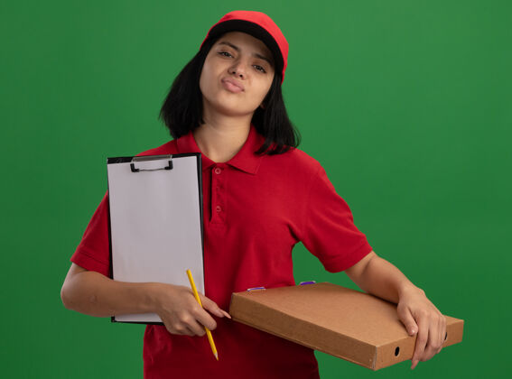 站着身穿红色制服 头戴鸭舌帽 拿着披萨盒和铅笔的年轻送货员站在绿色的墙上 表情自信帽子制服剪贴板