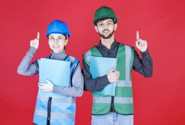 员工戴着头盔的男女工程师拿着蓝色文件夹 指着周围的人制服检查表姿势