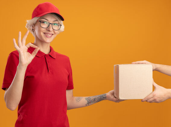帽子身着红色制服 头戴鸭舌帽的年轻女送货员站在橙色的墙上 友好地展示着收包裹的ok标志包装同时送货