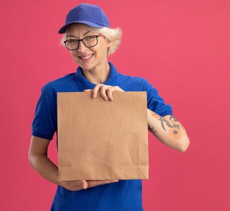 戴着身穿蓝色制服 戴着眼镜 拿着纸包的年轻送货员在粉红色的墙上欢快地微笑着制服年轻送货