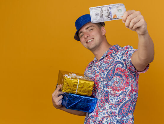 拿着微笑着的年轻人戴着蓝色帽子 拿着礼盒 拿着现金 站在橙色背景的相机前 留着复印空间戴着年轻人相机