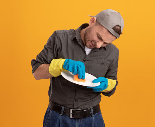 男人年轻的清洁工穿着休闲服 戴着帽子 戴着橡胶手套 拿着盘子和海绵洗盘子 站在橙色的墙上 脸上带着恼怒的表情海绵手套橡胶