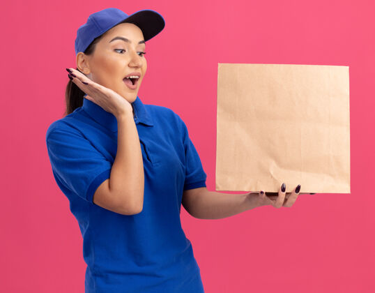 年轻年轻的送货员身穿蓝色制服 头戴鸭舌帽 手里拿着纸包裹 站在粉色的墙上 惊讶地看着它帽子包装交货