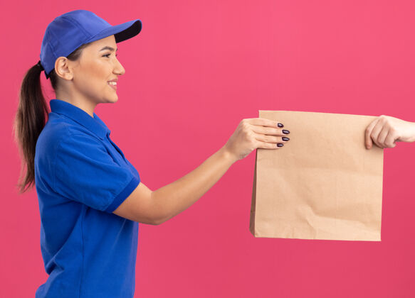 制服身着蓝色制服 头戴帽子的年轻送货员站在粉色的墙上 微笑着向顾客赠送纸质包装纸微笑女人