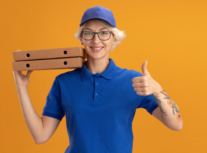 拿着穿着蓝色制服 戴着帽子 戴着眼镜 拿着披萨盒 微笑着在橙色的墙上竖起大拇指的快乐的年轻女送货员展示眼镜披萨