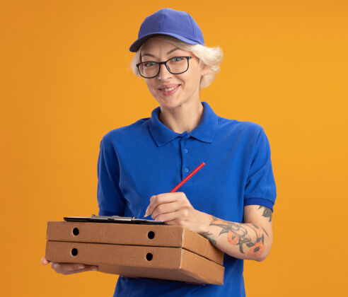 剪贴板身穿蓝色制服 戴着眼镜 拿着披萨盒和空白页的剪贴板 用铅笔在橙色墙上微笑着写着什么的年轻女送货员微笑铅笔披萨