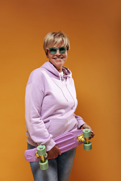 衬衫一位积极的老太太 金发 戴着时髦的太阳镜 穿着粉色的时髦运动衫和酷牛仔裤 微笑着拿着滑板牛仔年龄女