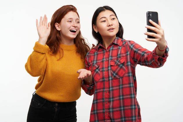 休闲人与生活理念两个可爱的女孩穿着黄色毛衣和格子衬衫女孩用视频电话向父母介绍她的朋友自拍隔着白墙孤立地站着模特在一起毛衣