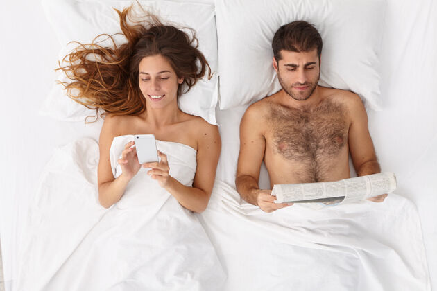 帅哥一家人睡在舒适的床上 女人用手机上网聊天 上网 沉迷于现代科技 男人看报纸 不说话应用程序床上用品在线