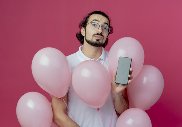 帅哥看着镜头帅哥戴着眼镜拿着气球和手机隔离在粉色背景上电话气球戴着