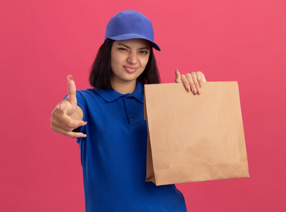 制服穿着蓝色制服 戴着帽子的年轻送货女孩拿着纸包 自信地用食指指着你站在粉色的墙上交货帽子年轻