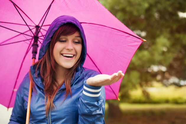 肖像快乐的女人在粉红色的雨伞下检查雨水站公园气象