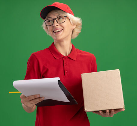 交付快乐的年轻送货员 身穿红色制服 戴着帽子 戴着眼镜 带着硬纸盒 拿着空白纸和铅笔的剪贴板 面带微笑地站在绿色的墙上 望着一旁年轻铅笔制服
