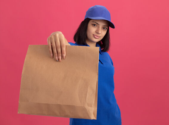 包装身穿蓝色制服 戴着帽子的年轻送货女孩站在粉红色的墙上 脸上带着自信的微笑 手里拿着纸包女孩举行交货