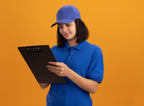 微笑身穿蓝色制服 戴着帽子的年轻送货员站在橙色的墙上 面带微笑地看着它帽子交货剪贴板