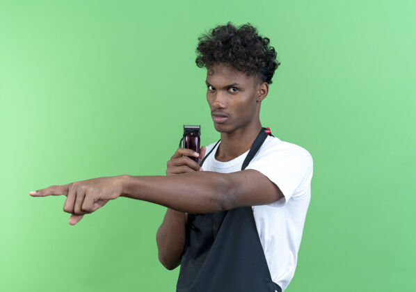 信心自信的年轻美国黑人男理发师穿着制服拿着理发器 指着绿色背景上孤立的一侧男剪子理发师