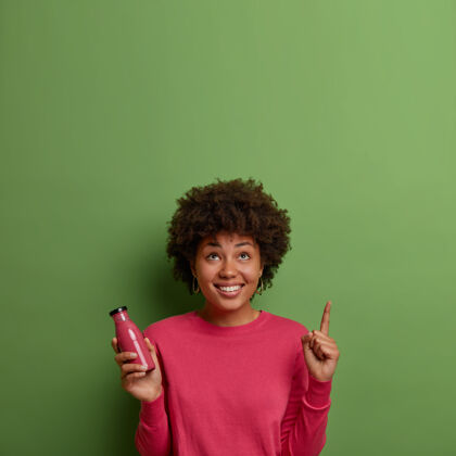 促销快乐的非洲裔美国妇女喝新鲜水果冰沙 用玻璃瓶盛着由超级食品制成的饮料 食指指向绿色墙壁上方 心情愉快健康的生活方式年轻有机牙关