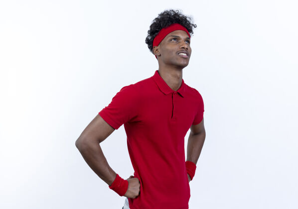 运动型抬头看着高兴的年轻的美国黑人运动男子戴着头带和腕带把双手放在臀部隔离在白色的背景年轻手腕带