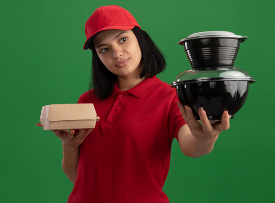 盒子身穿红色制服 头戴鸭舌帽 拿着食品包的年轻送货女孩站在绿色的墙上 看上去很自信帽子年轻递送