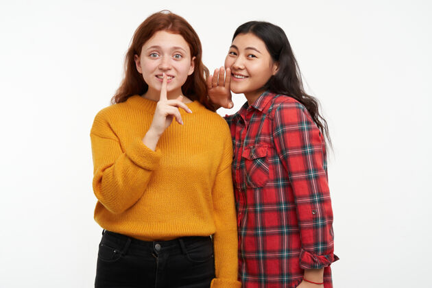 女性人和生活方式的概念两个时髦的朋友女孩低声对她的朋友 显示沉默的迹象穿着黄色毛衣和格子衬衫隔离在白色的墙壁耳语成人友谊