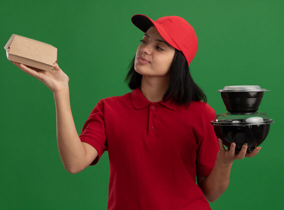 女孩身穿红色制服 头戴鸭舌帽 拿着食品包的年轻送货女孩站在绿色的墙上 看上去自信满面笑容帽子自信拿着