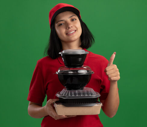 食物穿着红色制服 戴着帽子 手里拿着一叠食品包装 微笑着 友好地竖起大拇指 站在绿色的墙上 快乐的年轻送货女孩女孩年轻朋友