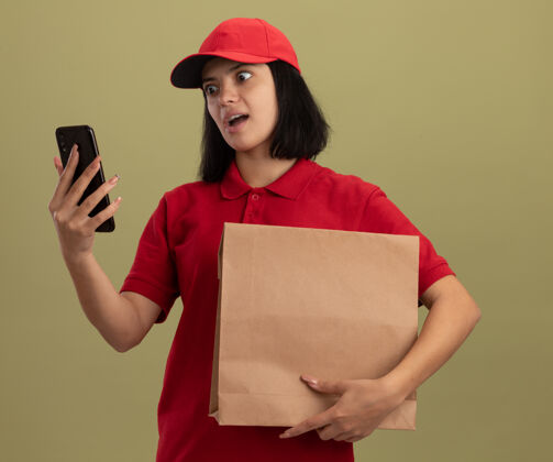 制服穿着红色制服 戴着帽子 拿着纸包的年轻送货女孩站在明亮的墙上 带着恼怒的表情看着她的智能手机持有恼火包装