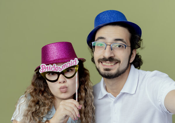 棍子高兴的年轻夫妇戴着粉红色和蓝色的帽子 女孩拿着化装眼罩 男孩拿着相机隔离在橄榄绿的背景上面具年轻橄榄