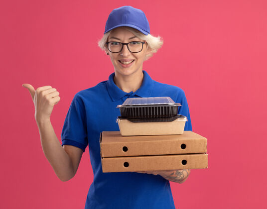 年轻人身穿蓝色制服 戴着眼镜 手持比萨饼盒和食品包的年轻送货员微笑着 用食指指着粉色墙壁的一侧食物制服女人