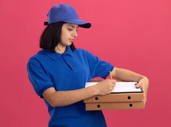 制服穿着蓝色制服 戴着帽子的年轻送货女孩拿着比萨饼盒和剪贴板 站在粉红色的墙上严肃地写着什么站着严肃的什么的