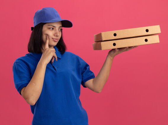 女孩穿着蓝色制服 戴着帽子 拿着披萨盒的年轻送货女孩站在粉红色的墙上沉思地看着披萨盒披萨表情帽子