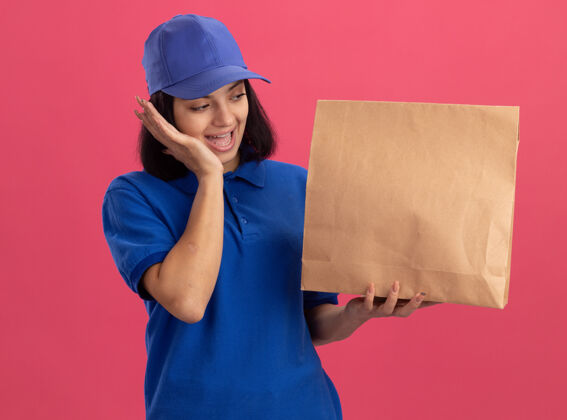 帽子身穿蓝色制服 头戴鸭舌帽的年轻送货员拿着纸包看着它 惊讶而快乐地站在粉色的墙上惊喜送货包装